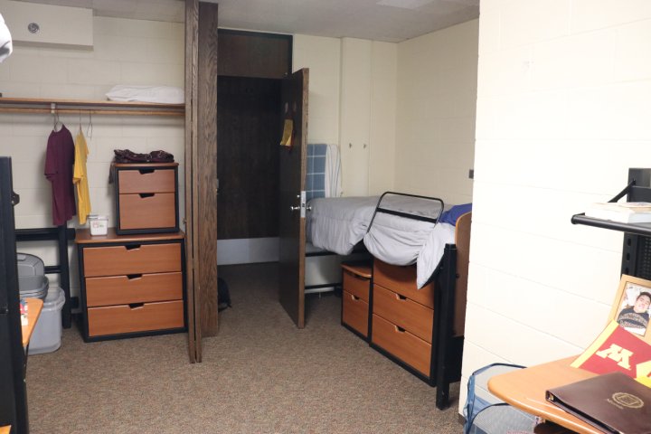 A college dorm room with a lofted bed, storage drawers, an open closet with hanging clothes, and a desk with a framed photo and books. Simple and organized layout.